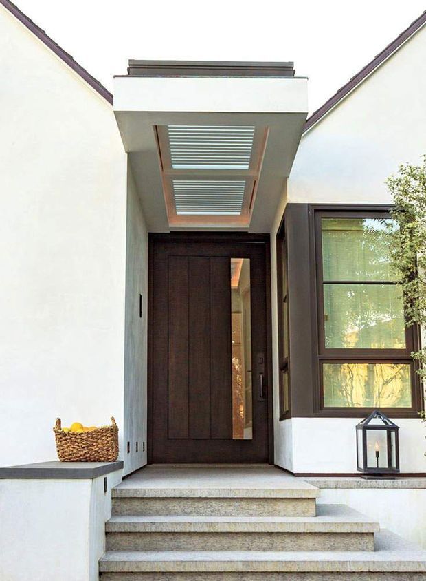 A close-up exterior view of a refined residential entryway. Broad pale rendered walls frame a dark timber front door with a narrow glazed sidelight, accessed by several light stone or concrete steps. A projecting flat-roof canopy contains two timber-framed slatted skylight panels, while dark-framed windows sit to the right. A built-in ledge with a woven basket and a small lantern adds understated detail.
