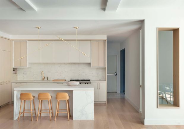 Bright contemporary open-plan kitchen with a white island and three timber stools, pale integrated cabinetry, a light stone-look splashback and benchtop, brass-toned fittings, and light timber flooring. A hallway extends to the right, with a full-height wall mirror reflecting a dining or living area. White ceiling beams and minimal pendant lights add subtle architectural detail.