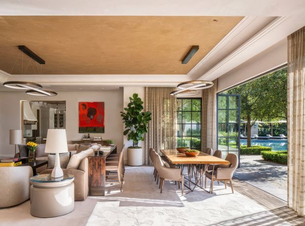 A refined open-plan living and dining area with a large dining table, upholstered dining chairs, sofas, occasional tables, and contemporary pendant lighting. Full-height glazed doors open to a landscaped outdoor area and swimming pool. The room features a warm wood-toned ceiling, pale walls, light flooring, extensive curtains, dark-framed glazing, and built-in architectural mouldings.