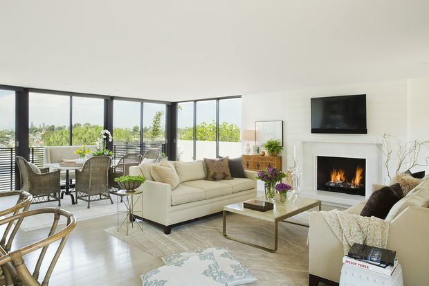 Bright, spacious open-plan living room with floor-to-ceiling corner glazing, a dining area, cream sofas, a glass-topped coffee table, wall-mounted television, and a contemporary fireplace with a visible fire. The room has pale timber flooring, white walls and ceiling, dark window frames, and abundant greenery visible outside.