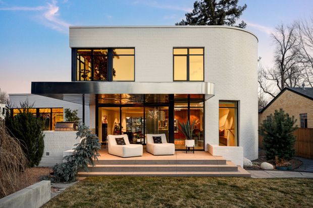 Photograph of a contemporary two-storey residential home at dusk, featuring a rounded white masonry upper volume, extensive black-framed glazing, a projecting flat-roofed terrace canopy, and warm interior lighting. The rear patio has broad steps, outdoor lounge chairs, built-in planters, and a landscaped suburban yard. A neighbouring pitched-roof house is visible to the side.