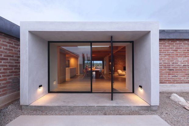 Front exterior view of a compact contemporary extension or pavilion, framed in smooth pale plaster/concrete with a large black-framed glazed opening and central pivot door. Exposed red brick boundary walls, warm timber ceiling lining, and a warmly lit interior are visible through the glazing. Gravel landscaping and discreet wall lights reinforce the minimalist architectural presentation.