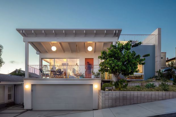 Front exterior view of a contemporary two-storey urban residence at dusk. The house features a broad projecting upper canopy with exposed underside beams, a sheltered balcony with metal railings, extensive upper-level glazing, a large integrated garage, and a raised landscaped frontage with a tiled retaining wall and mature tree. The palette is predominantly light grey, white, and muted timber tones, with warm interior and exterior lighting.