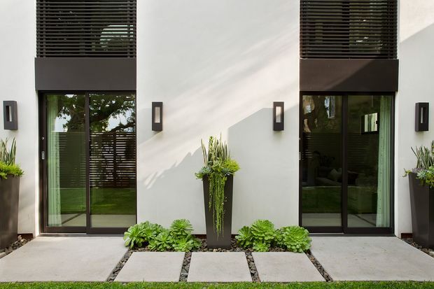 A symmetrical modern residential facade with white rendered walls, two tall dark-framed glazed entry doors, horizontal dark louvres above, and matching black wall lights. Slim charcoal planters and low succulents line the concrete-paver entry path, creating a restrained contemporary exterior.