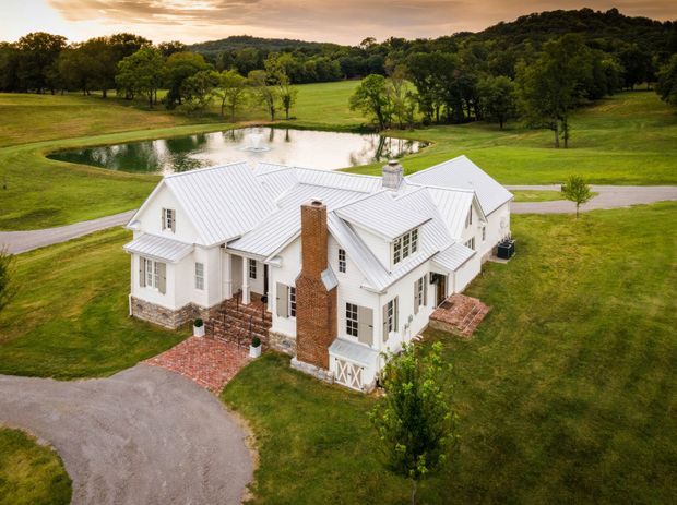 Aerial view of a large white country house set within expansive landscaped grounds. The home has white weatherboard-style walls, multiple steeply pitched gabled roof sections with standing-seam metal roofing, multi-pane windows, and a prominent red brick chimney. Brick-paved paths and terraces surround the house, with a pond, lawns, mature trees, and rolling rural landscape beyond.