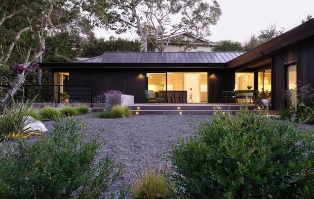 A low, single-storey contemporary residence set within a landscaped garden courtyard. The dark charcoal exterior has crisp horizontal volumes, a low standing-seam metal roof, extensive warm-lit glazing and timber-lined openings. Gravel paving, native-style planting, and subtle step lighting create a secluded evening garden setting.