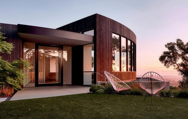 A contemporary single-storey home photographed at dusk, featuring a dramatic curved timber-clad facade, extensive floor-to-ceiling glazing, and a projecting flat-roofed entry canopy. The house opens onto a manicured lawn with two sculptural outdoor lounge chairs, landscaped planting, and expansive distant views.
