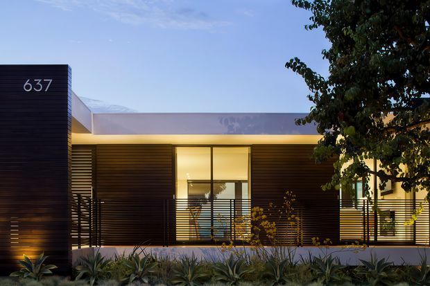 A contemporary single-storey residential facade photographed at dusk, with a dark horizontal slatted entry volume marked 637, long low rooflines, expansive illuminated glazing, and a projecting covered walkway screened by fine horizontal metal railings. Layered agave-like planting and a mature tree frame the front elevation.