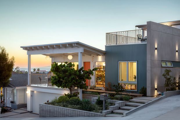 Exterior view of a contemporary multi-level residence on a sloping urban site at dusk. The home features a flat roof, projecting covered terrace with exposed roof beams, blue-grey vertical cladding, smooth dark grey wall panels, extensive outdoor stairs and terraces, rooftop/balcony railings, and warmly lit glazing. Landscaped garden beds and neighbouring rooftops are visible.