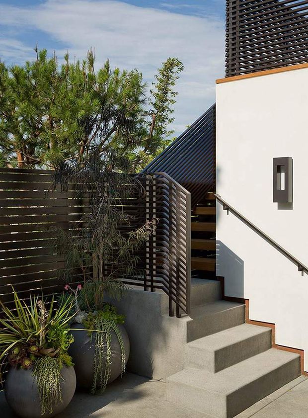 Exterior view of a contemporary residential entry, featuring a concrete stairway with dark horizontal slatted metal or timber screening, a white rendered wall, simple modern wall light, and landscaped planter pots with established greenery. The layered privacy screens and geometric stair composition create a refined, modern facade.