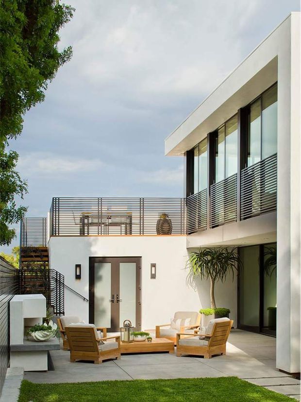 A bright contemporary two-storey courtyard residence with white rendered walls, extensive dark-framed glazing, horizontal metal privacy railings, and a flat roof. The landscaped outdoor living area includes a paved patio, timber lounge furniture, a low coffee table, built-in outdoor fireplace or bench element, potted plants, and a small lawn. An upper terrace and exterior stair are visible.