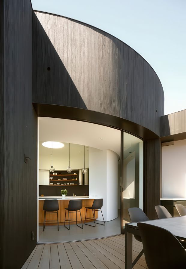 A contemporary curved architectural facade clad in dark vertically ribbed panels wraps around a glazed opening to a refined kitchen and dining area. The view is from a timber-decked outdoor space, looking through large curved glazing at a kitchen island with bar stools, pendant lights, pale cabinetry, and a dark feature shelving wall. Strong geometric curves, contrasting dark cladding, warm timber, and abundant glazing define the composition.
