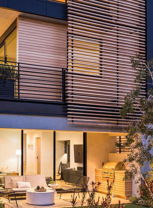 A dusk exterior view of a contemporary multi-level residence with dark painted facade elements, warm timber horizontal cladding, and a prominent timber slatted privacy screen across the upper storey. Black metal balcony railings, large glazed openings, and a sheltered ground-floor outdoor living area with lounge seating and a built-in barbecue are visible. Warm interior lighting creates an inviting atmosphere, while planted landscaping frames the foreground.