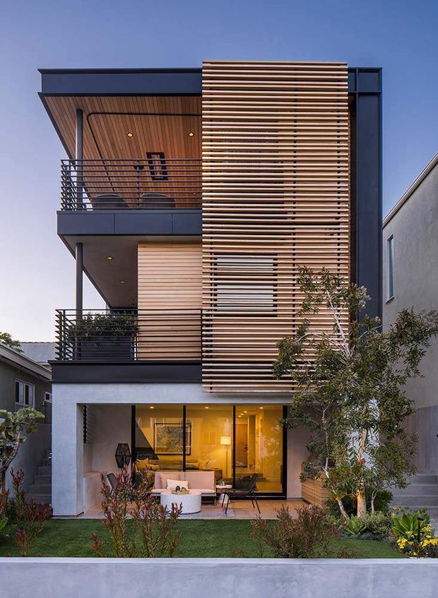 Multi-storey contemporary urban residence viewed from the front, with a dark flat-roofed facade, projecting balconies, extensive glazing, and a prominent vertical screen of horizontal timber slats. The upper balcony has a timber-lined ceiling and dark metal balustrades, while the ground level opens to a furnished living area and landscaped garden.