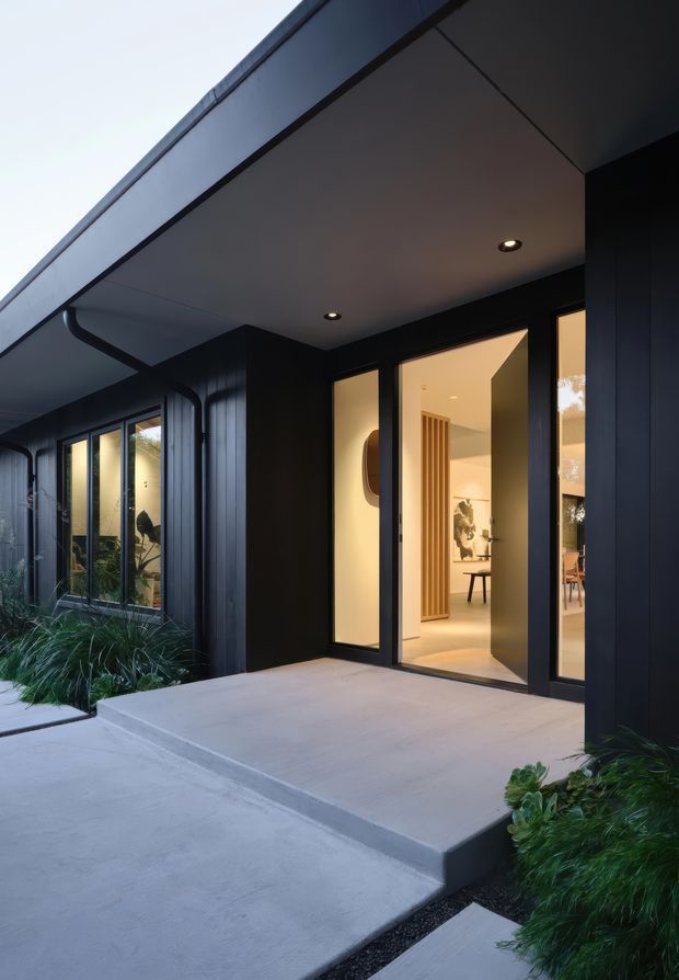 Exterior dusk view of a contemporary home entry, with a deep covered porch, dark vertical cladding, black-framed full-height glazing and a broad concrete entrance slab. Warm interior lighting is visible through the open glass door, while low ornamental planting borders the concrete pathway.