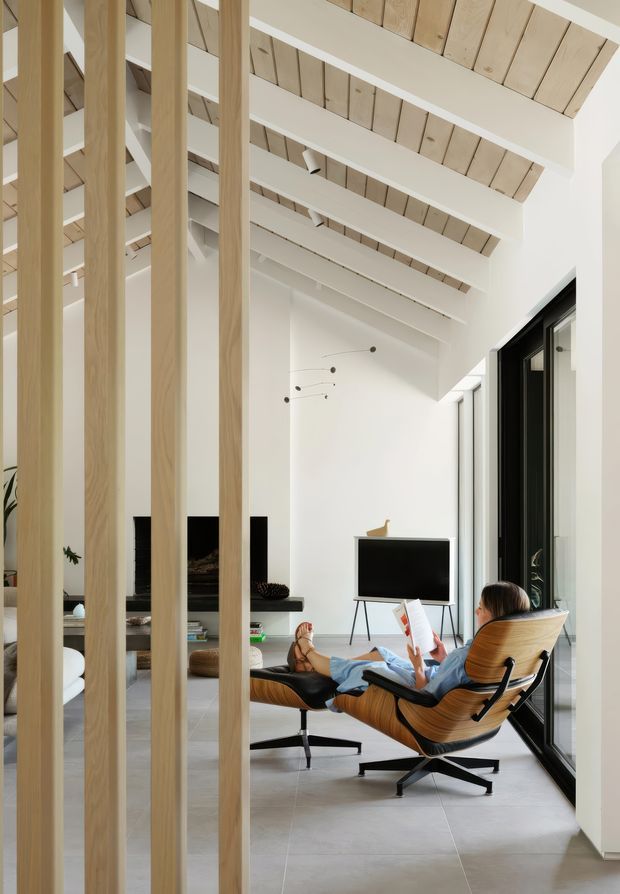 A bright, double-height contemporary living room viewed through vertical light timber screening battens. The room features exposed timber roof boards and white structural beams, pale tiled flooring, extensive dark-framed glazing, a black fireplace surround, a television, and a sculptural leather lounge chair with ottoman. A linear mobile light hangs in the open space, creating an airy, refined interior with strong indoor-outdoor connection.