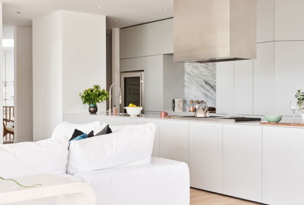 Bright open-plan contemporary kitchen viewed from the adjoining living area. The kitchen features extensive handleless pale cabinetry, a long island or peninsula with a light stone benchtop, a large marble-look splashback, integrated appliances, and a box-shaped rangehood. A white sofa occupies the foreground, with a glimpse of dining furniture at left. The palette is predominantly white and warm pale neutrals, creating a clean, refined atmosphere.