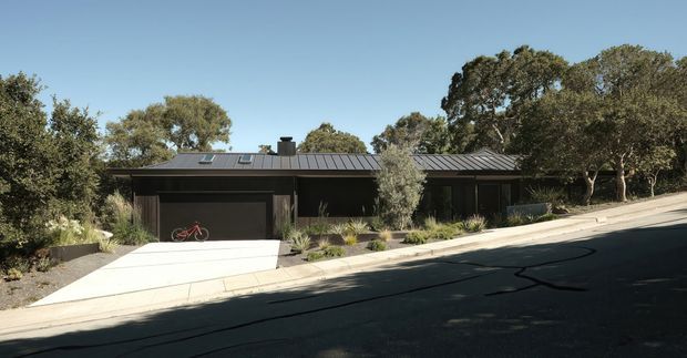 A low, single-storey dark-clad residence viewed from across a sloping suburban street. The house has a long horizontal composition, a dark standing-seam pitched roof, broad dark garage and entry openings, and restrained native-style landscaping with mature trees. A concrete driveway and sloped footpath lead up from the street.