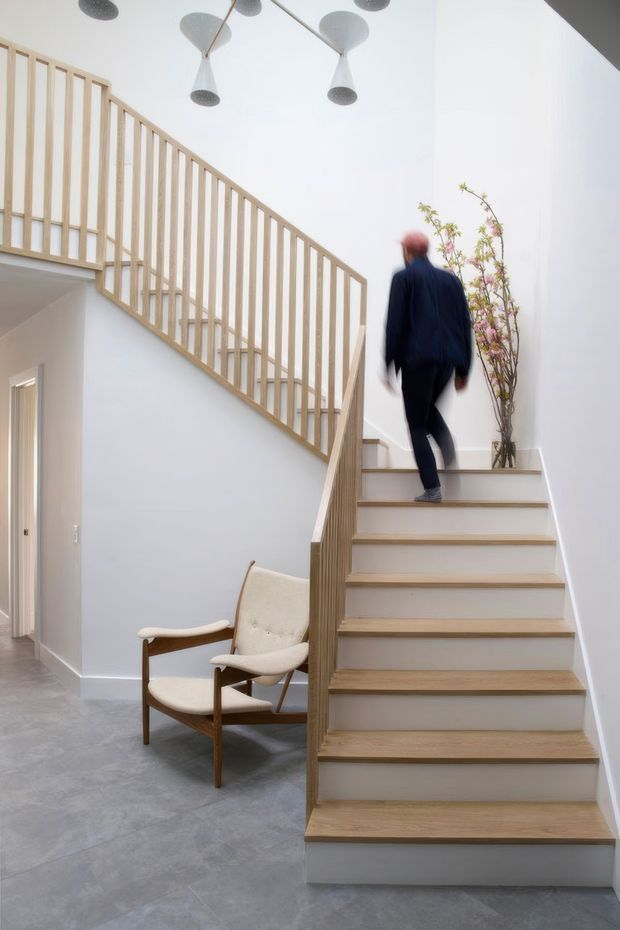 Bright contemporary interior stairway with a light timber staircase and slim vertical timber balustrade rising through a tall white stair hall. The stair treads and handrail contrast with pale grey polished flooring and white walls. Sculptural pendant lights hang above, while a mid-century style upholstered lounge chair and a tall indoor plant sit beneath the stairs. A blurred person is walking up the stairs.