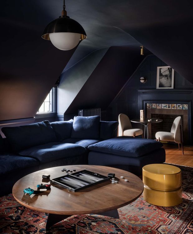 A dark, atmospheric living room with a dramatic sloped ceiling and deep navy walls. A large navy sectional sofa and ottoman face a round timber coffee table, with a patterned Persian-style rug beneath. Pendant lights, a compact round side table, sculptural cream armchairs, a framed artwork, and a dark fireplace or media surround complete the richly layered interior.