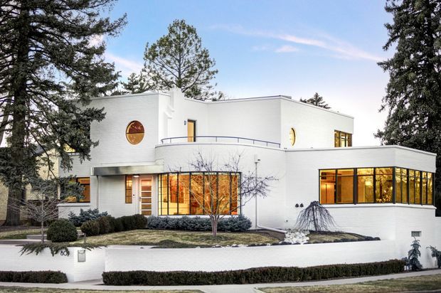 Exterior view of a large white modernist residence set among mature trees and landscaped grounds. The house has strongly geometric cubic forms, a flat roof, curved balcony and projecting glazed living spaces with warm interior lighting. Distinctive round window openings and dark metal balcony railings add Art Deco-influenced detailing.