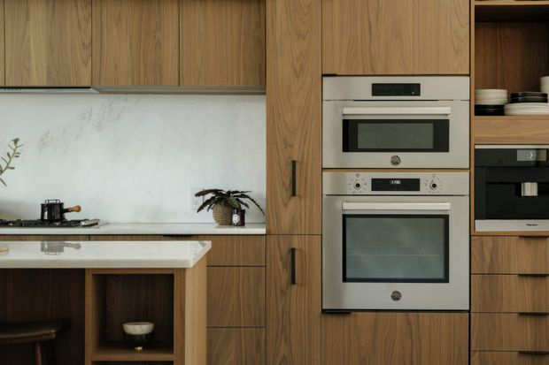A refined contemporary kitchen with warm timber cabinetry, integrated stainless-steel double ovens, a pale veined stone splashback and benchtop, and a compact island or peninsula with open timber shelving and seating. Dark hardware and appliances provide contrast, while open shelving and plants add understated warmth.