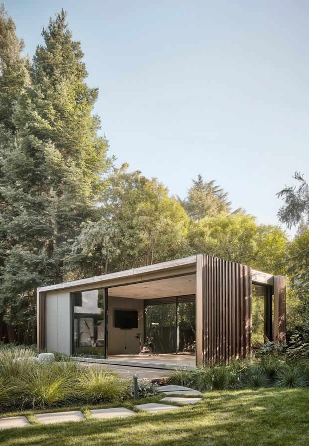 A compact modern pavilion set within a lush landscaped garden. The single-storey structure has a flat roof, extensive floor-to-ceiling glazing, dark vertically ribbed exterior cladding, warm timber soffits, and open sliding glass walls. A simple stepping-stone path crosses the lawn toward the pavilion, which contains a small exercise area visible through the glazing.