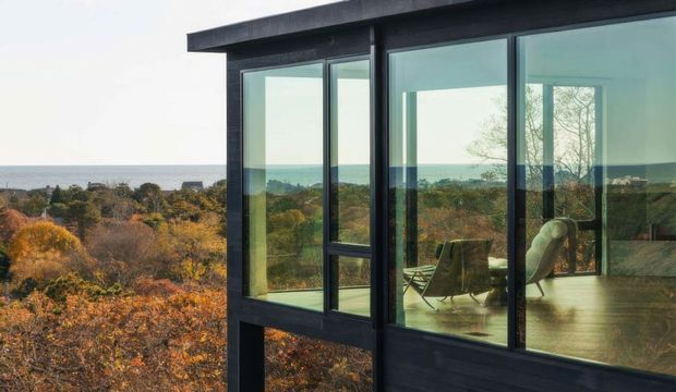 Exterior view of a contemporary dark-clad building with extensive floor-to-ceiling glazing, overlooking a wooded landscape and distant water. Through the windows, a simply furnished interior with timber flooring and lounge seating is visible. The image emphasizes the strong connection between the glass facade and the surrounding natural setting.