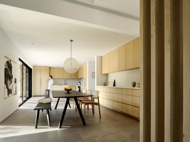 A bright, open-plan dining and kitchen interior viewed from a timber-framed entry or hallway. The room features extensive pale timber cabinetry, a long built-in sideboard, a dark dining table with mixed timber chairs and bench seating, large black-framed glazed doors, a sculptural pendant light, and large-format neutral floor tiles. The restrained palette and clean built-in joinery create a warm contemporary atmosphere.