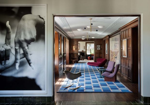 View through a wide doorway into a richly detailed, wood-panelled interior resembling a formal living room or sitting room. The room features dark timber built-in cabinetry and wall panelling, ornate white ceiling mouldings, multiple modern pendant and ceiling lights, a blue geometric area rug over dark timber flooring, and a mix of mid-century lounge chairs and a low sofa. A large monochrome artwork and dark green tiled wainscot frame the foreground entry.