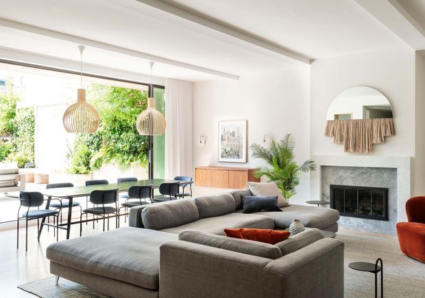 Bright open-plan living and dining area with a large grey sectional sofa, a dining table beside full-height glazing, a contemporary fireplace, timber sideboard, indoor palm, woven pendant lights, and a round wall mirror with textile decor. The space has white walls and ceiling beams, a light neutral palette, and strong indoor-outdoor visual connection to greenery.
