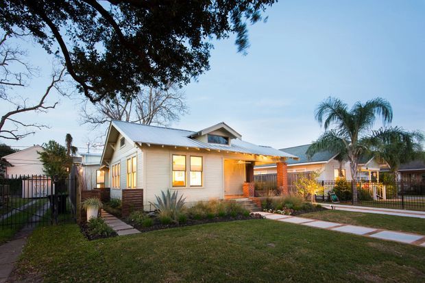 Single-storey suburban residence photographed at dusk, featuring a light weatherboard-clad main house with a pitched gabled roof, projecting entry porch, warm illuminated windows, brick columns, landscaped front garden, and concrete stepping paths. Mature trees, palms, fencing, and neighbouring homes surround the property.