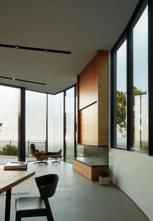 A contemporary open-plan living space with expansive floor-to-ceiling glazing, a sculptural corner window, polished concrete flooring, and a warm timber feature wall with integrated cabinetry and a dark stone-like bench. The room includes a lounge chair and ottoman, dining or work seating in the foreground, minimalist linear recessed lighting, and views toward a balcony and landscape beyond. The composition is bright, refined, and strongly connected to the outdoors.