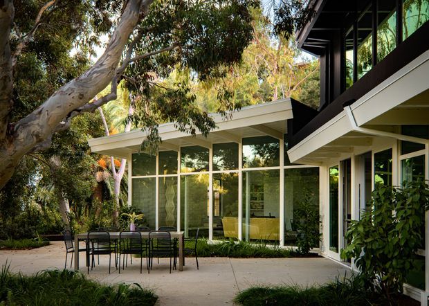 Exterior view of a contemporary single-storey home set within established eucalyptus-like garden planting. The image focuses on broad floor-to-ceiling glazed walls, projecting low-pitched roof forms, dark structural framing, and a paved outdoor dining terrace. Interior furnishings are visible through the glazing, while mature trees and landscaping surround the building.