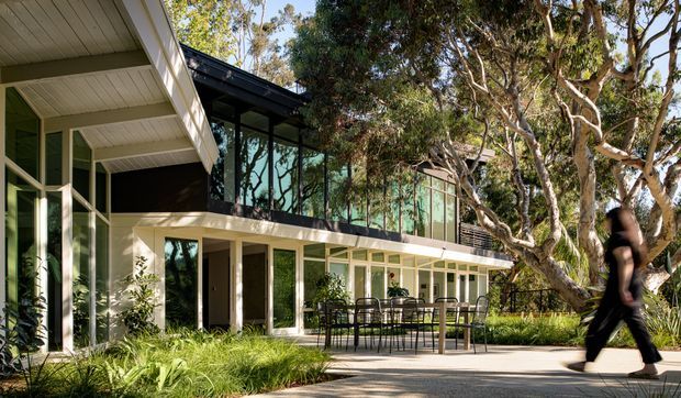 A contemporary multi-level residential building set among mature eucalyptus trees and landscaped planting. The facade combines extensive floor-to-ceiling glazing with slender dark frames, pale rendered or painted walls, and projecting roof planes. A covered outdoor terrace with a dining table and chairs sits beside a paved walkway, while a blurred person provides scale.