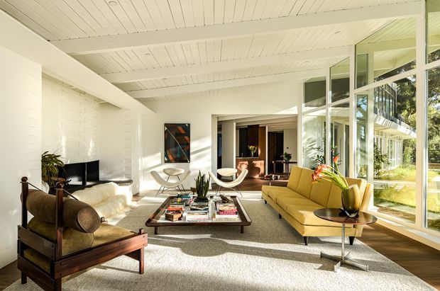 Bright, spacious mid-century-inspired living room with white painted brick walls, a white boarded ceiling with exposed beams, expansive floor-to-ceiling glazing, and warm timber flooring. The room contains a large cream rug, vintage-style leather and timber seating, a mustard-yellow sofa, sculptural lounge chairs, plants, and a low coffee table. Strong indoor-outdoor connection through the glazed wall, with a secondary sitting or dining area visible beyond.