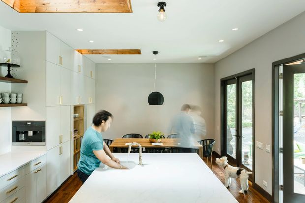 Bright, contemporary open-plan kitchen and dining area. The foreground features a large white island with an integrated sink and brushed metal tap, backed by full-height white cabinetry with brass-toned hardware and integrated appliances. A timber dining table with black chairs sits beside dark-framed glazed doors, while exposed timber ceiling beams and warm timber flooring add contrast. The room has a clean, airy feel with pendant and recessed lighting.