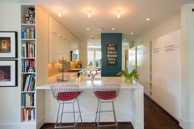 Bright, narrow galley-style kitchen viewed from the living area, with a large white island and two wire bar stools in the foreground. Floor-to-ceiling white cabinetry runs along both sides, with integrated appliances, a warm mosaic-tile backsplash, pale benchtops, dark timber flooring, open shelving filled with books, and a teal feature panel with decorative lettering. Recessed and pendant-style ceiling lights create a clean, inviting contemporary interior.
