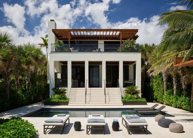 A contemporary two-storey residence viewed from the pool courtyard. The white rendered facade has tall dark-framed openings, a glass-balustraded upper balcony, and a timber pergola with exposed slats over the upper level. A swimming pool, paved pool terrace, loungers, outdoor seating, palms and dense tropical landscaping occupy the foreground and sides.