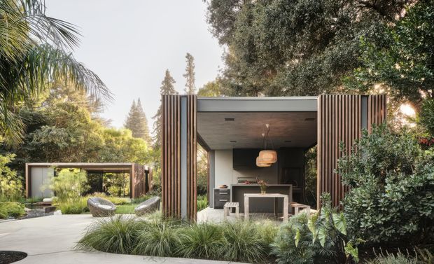 A contemporary garden pavilion or outdoor entertaining room set within dense landscaped greenery. The compact structure has a flat roof, deep open-fronted glazing or doors, vertical timber screening at both sides, and a dark neutral interior with a compact kitchen or bar area and central island. A paved terrace with outdoor lounge seating and ornamental grasses surrounds the pavilion.