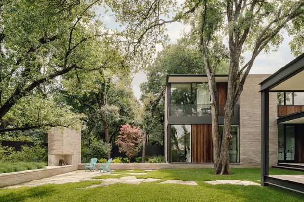 A contemporary multi-level residential building set within a mature, leafy landscape. The composition shows a dark brick and timber-clad facade, extensive floor-to-ceiling glazing, projecting flat roof forms, and a paved garden courtyard with stepping stones and outdoor seating. Large established trees integrate the architecture with the garden setting.