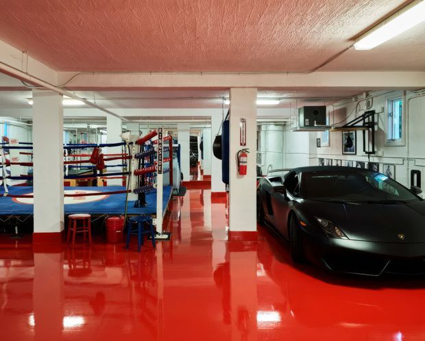 A large basement-style boxing gym and vehicle storage space with a glossy red floor and red textured ceiling. Boxing rings, heavy bags, training equipment, mirrors, exposed services, fluorescent lighting, and a black sports car occupy the open interior. White structural columns and walls create a stark high-contrast industrial training environment.
