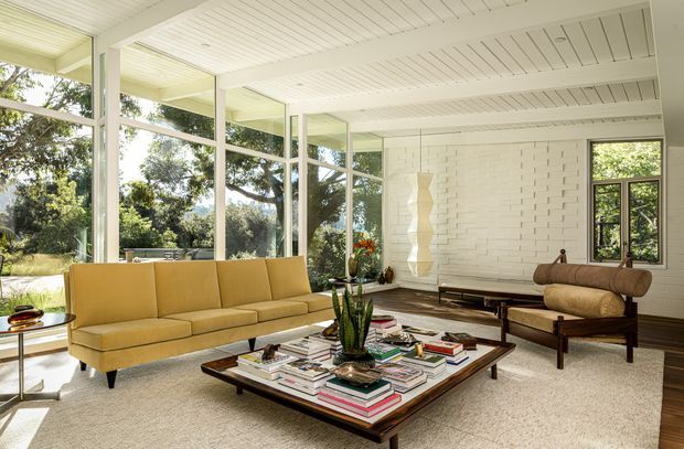 Bright mid-century-inspired living room with expansive floor-to-ceiling glazing, white-painted brick walls, a white slatted ceiling, and warm timber furnishings. A mustard-yellow sofa and sculptural lounge chair surround a low rectangular coffee table layered with books and decorative objects. Lush greenery is visible outside through the windows, creating an airy indoor-outdoor connection.