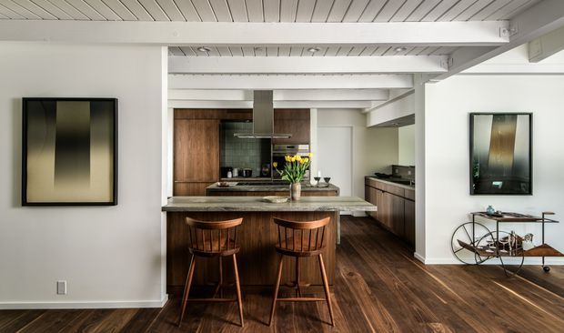 Open-plan kitchen and living area with a prominent central island, two wooden bar stools, dark timber cabinetry, stone benchtops, and rich timber flooring. White-painted exposed ceiling beams and lining create a bright, structured interior, while framed artwork and a compact console add restrained mid-century character.