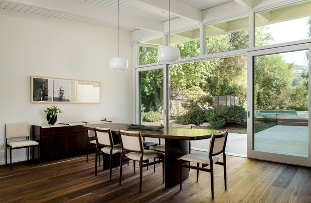 A bright, refined dining room with a long dark timber dining table and upholstered dining chairs, positioned beside expansive floor-to-ceiling glazing and a glazed door opening to a landscaped outdoor area. The room features dark timber flooring, a white painted slatted ceiling with exposed beams, a dark timber sideboard, minimalist framed wall art, and sculptural pendant lights. Lush greenery and a pool or water feature are visible outside.