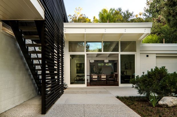 Exterior view of a modern residential extension or courtyard connection. A pale rendered or painted masonry wall and white horizontal-clad flat-roofed pavilion frame full-height glazed doors and clerestory windows opening to a dining area. A black louvred privacy screen and external stair form a strong vertical architectural element on the left, while a textured concrete path and landscaped planting occupy the foreground.