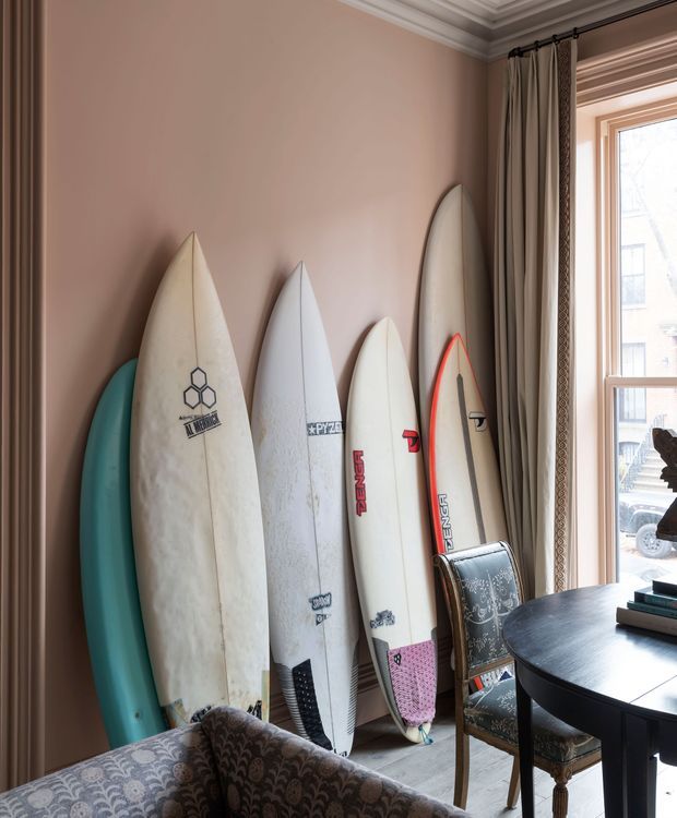 A warmly styled traditional interior corner with several surfboards displayed upright against a dusty rose wall beside a tall window. Decorative wall and ceiling mouldings, patterned curtains, an upholstered patterned chair and sofa edge, and a dark round table create a refined, eclectic room setting.