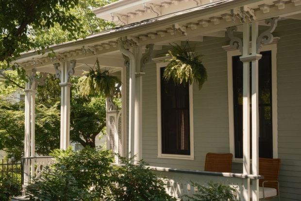 A close exterior view of a heritage-style veranda with pale green weatherboard cladding, ornate white timber posts and brackets, decorative fretwork, a projecting eave, tall dark-framed windows, and hanging ferns. Lush garden foliage partially screens the veranda, creating a shaded and characterful residential facade.