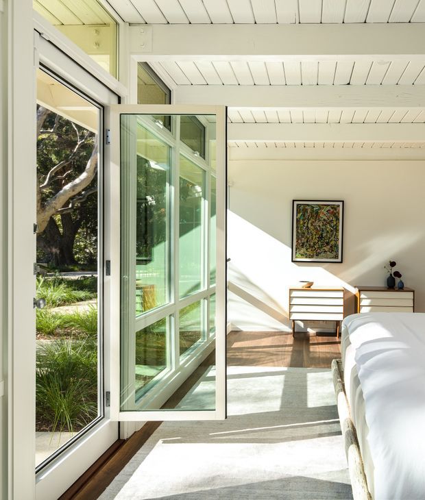 A bright bedroom photographed from the threshold of open glazed doors, with a white-painted timber-lined ceiling, pale walls, warm timber flooring, a light area rug, and a white bed in the foreground. Mid-century-inspired timber dressers sit beneath a colourful framed artwork, while abundant glazing opens toward a landscaped garden. Strong sunlight creates diagonal shadows across the room.