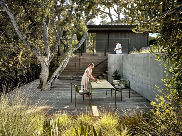 A landscaped outdoor entertaining terrace set within mature planting, with a timber deck, outdoor dining table and chairs, a built-in concrete retaining wall, dark metal balustrades, and a raised upper deck adjoining a dark-clad contemporary building. Ornamental grasses and established trees create a private, natural setting.